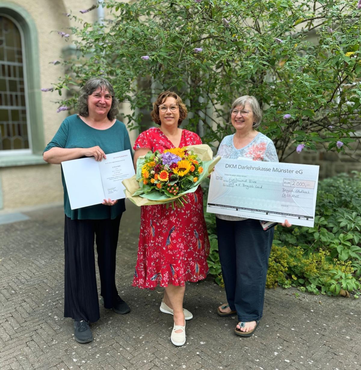 Links im Bild stehen Dorothee van den Borre, Melanie Müller-Spahn und Dr. Birgit Timmermann. Sie lächeln in die Kamera. In den Händen halten sie einen Blumenstrauß, eine Urkunde und einen symbolischen Scheck über 2.000 Euro.
