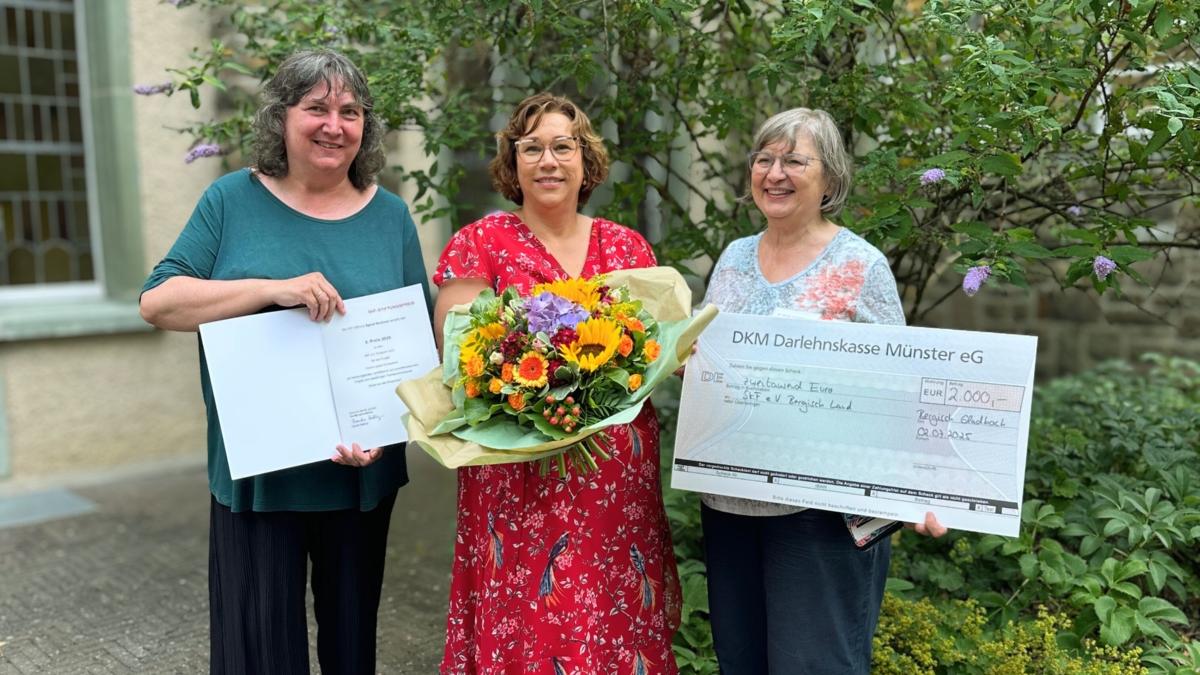 Links im Bild stehen Dorothee van den Borre, Melanie Müller-Spahn und Dr. Birgit Timmermann. Sie lächeln in die Kamera. In den Händen halten sie einen Blumenstrauß, eine Urkunde und einen symbolischen Scheck über 2.000 Euro.